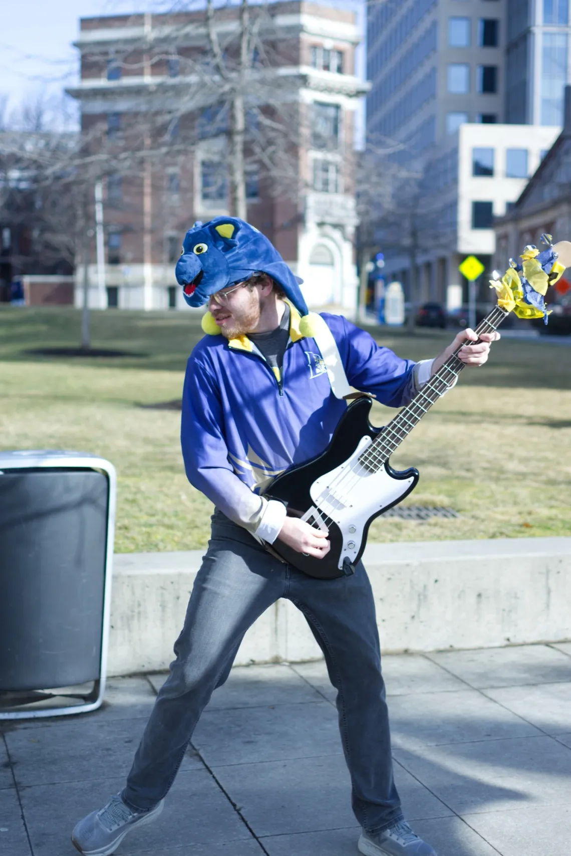 A man in purple and yellow attire posing with an electric bass in a public square.