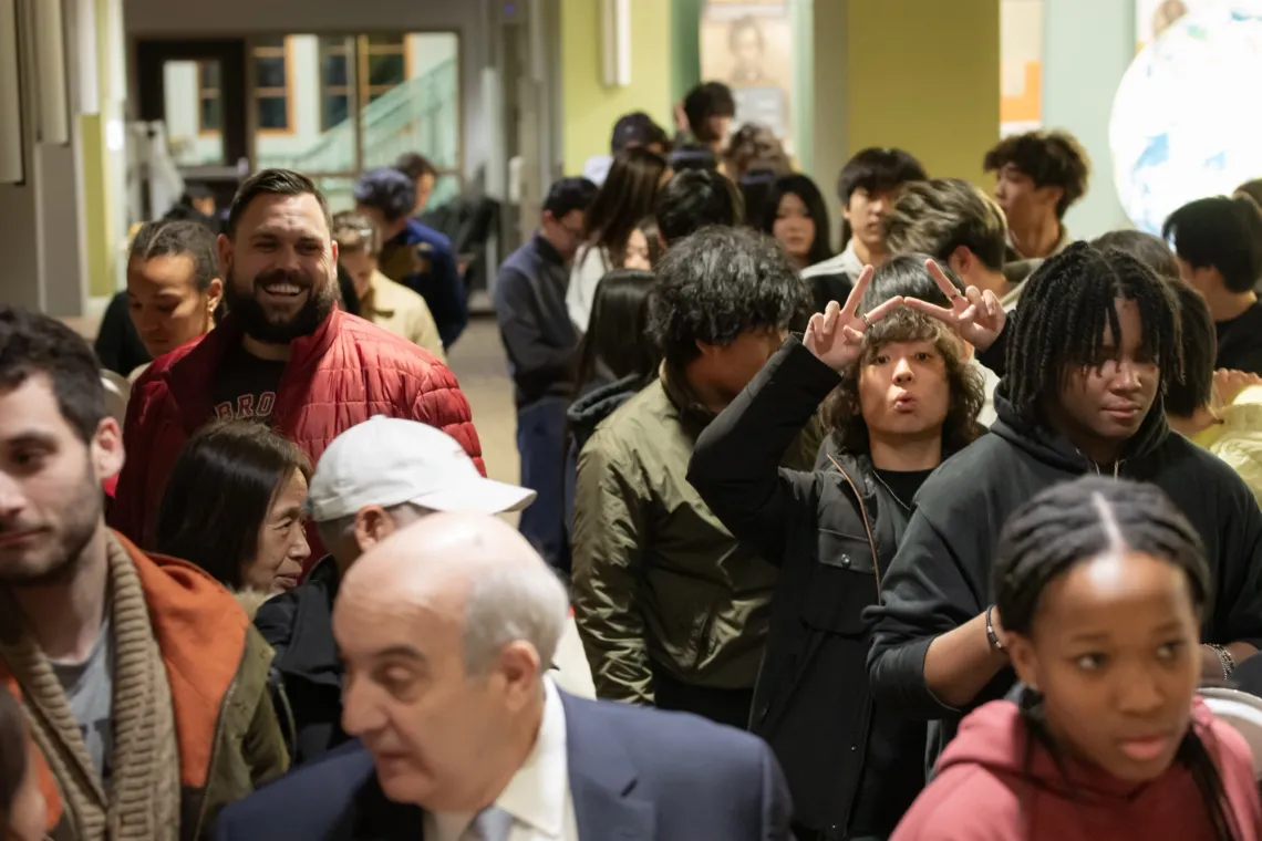 In a crowd, an event participant, in black attire, holds up two peace signs with his hands to pose for the camera.