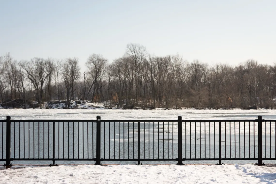 Black railing separates a snow-covered overlook and an icy river. Winter trees make up the background.