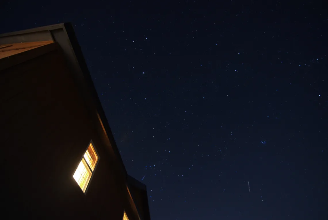 Dots of stars are scattered across a dark sky. In the foreground, a building with golden light leaks out of the windows.