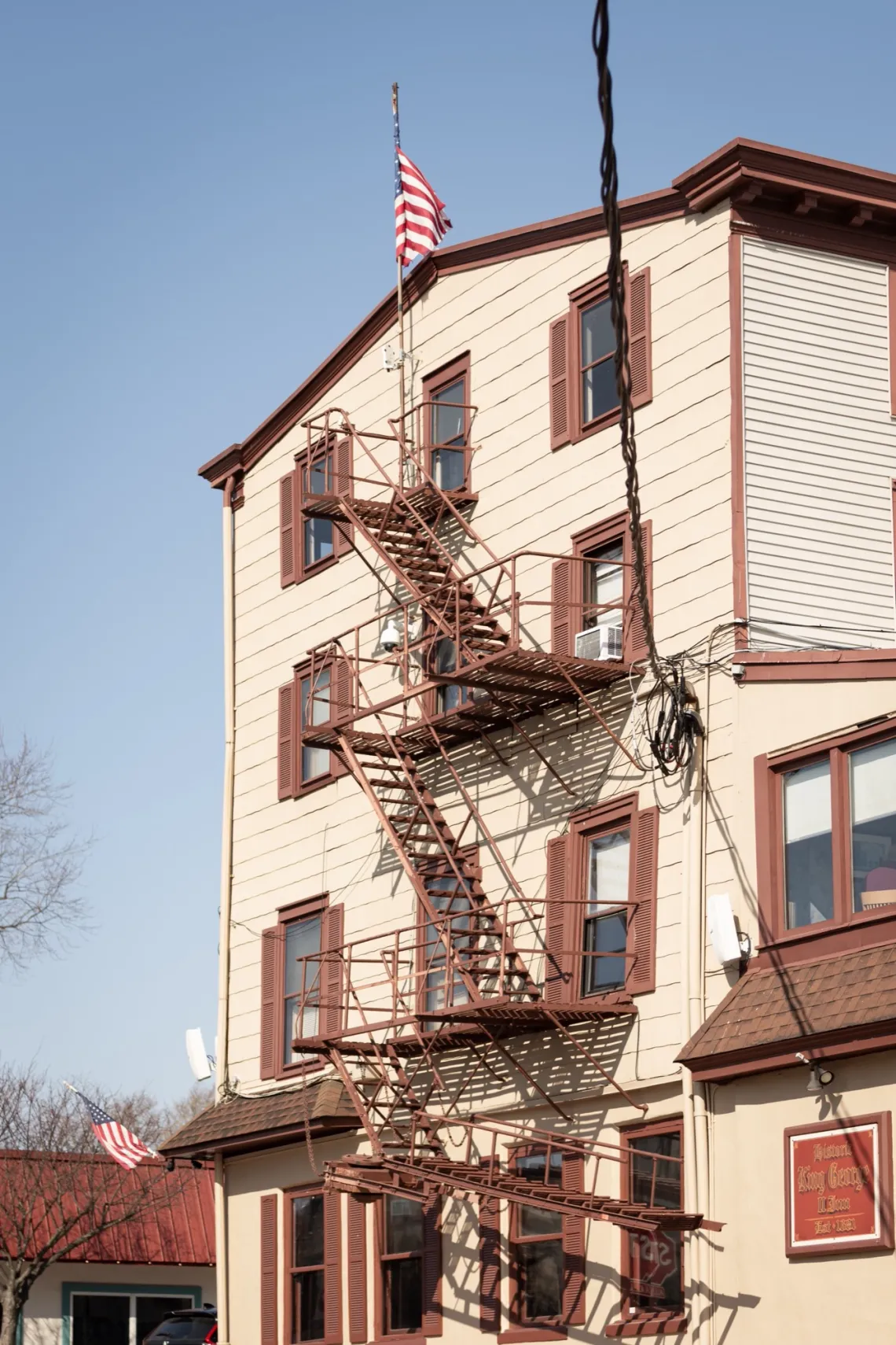 On the side of a building, a set of exterior stairs, with railings, leads almost the entire way to the ground, stopping short. A flag of the United States waves above.