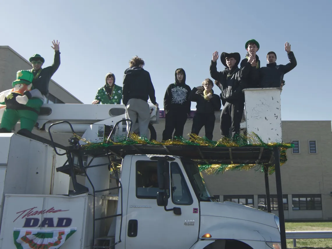 A group of people wave from on top of a truck. The truck is dressed with green Saint Patrick's Day decorations.