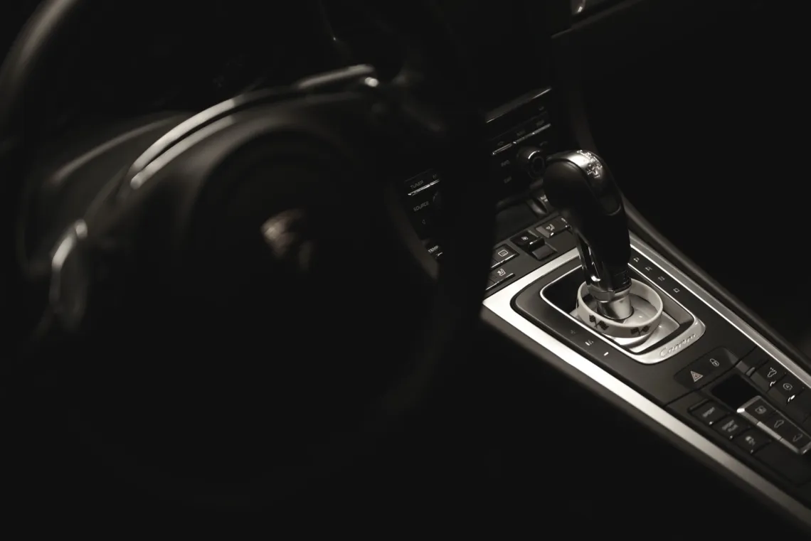 The dark interiors of a Porsche car. The Porsche logo, left, is out-of-focus, while silver accents of the transmission and button controls, right, include a white wristband of racing flags.