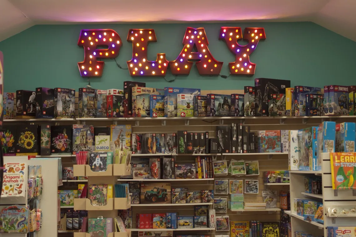 Shelves of children's books and LEGO sets are displayed in a toy store, under a big, colored, light-up sign reading "PLAY".