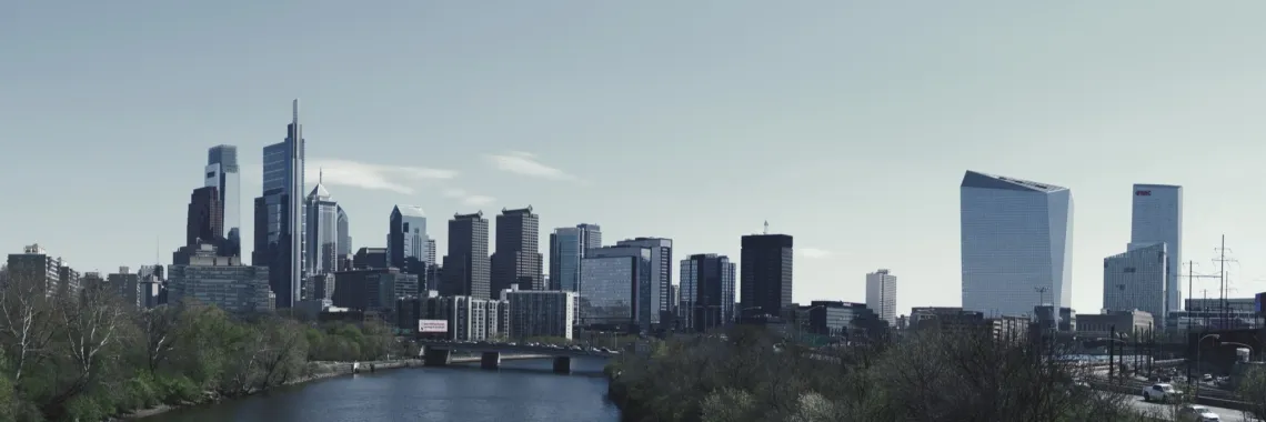 A row of skyscrapers behind a river and bridge in Philadelphia, framed by trees (left) and a highway (right)