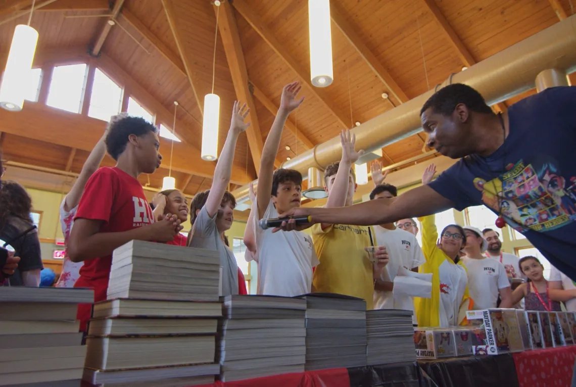 A man in a blue shirt, right, leans over a table full of books and figurines, passing a microphone to a boy, left, with face paint. Several people are raising hands.