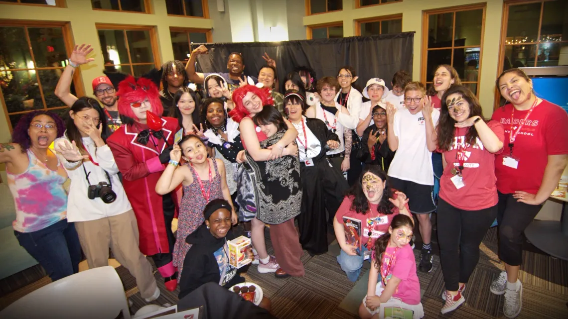 At a convention, many people in cosplay and colorful attire gather in front of a black curtain to cheer and wave at the camera.