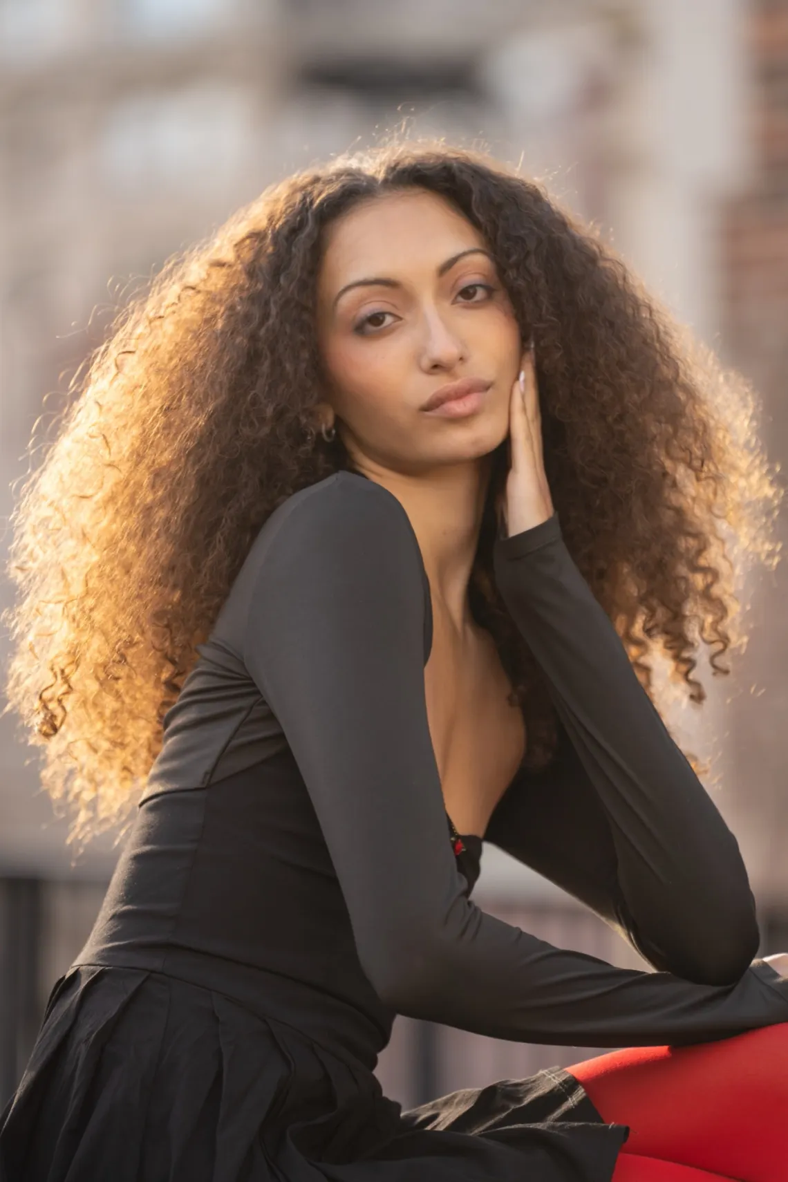 A woman in gray and red attire is lit by the setting sun, in front of an urban background.