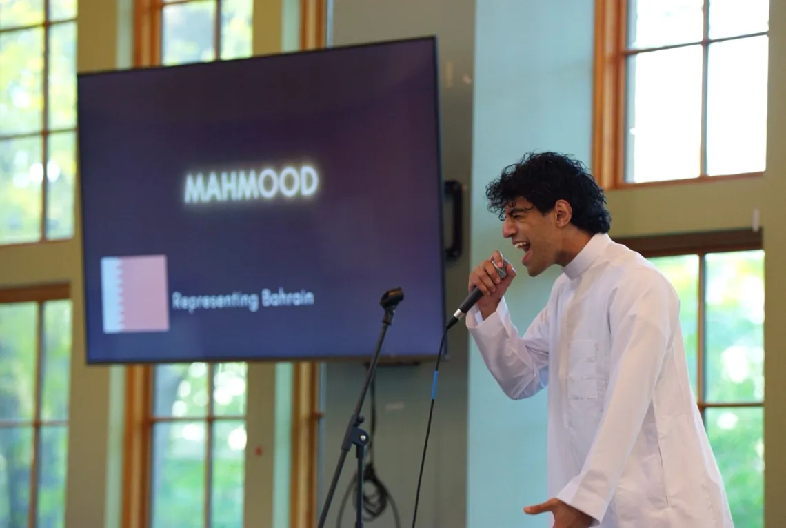 Man (right) in white attire sings into a wired microphone. The screen behind him (left) reads "MAHMOOD Representing Bahrain" with a white and red Bahraini flag.