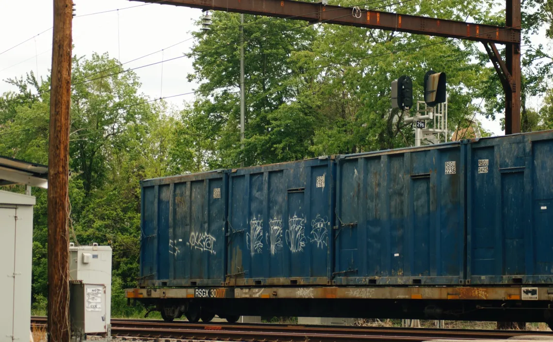 A blue train boxcar is the last to pass by this train crossing, surrounded by green trees and wires.