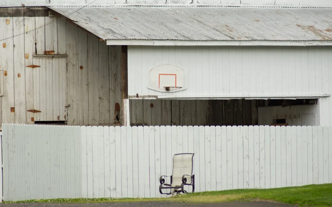 White chair is in front of a white fence, which surrounds a white barn with a white and red basketball net attached.