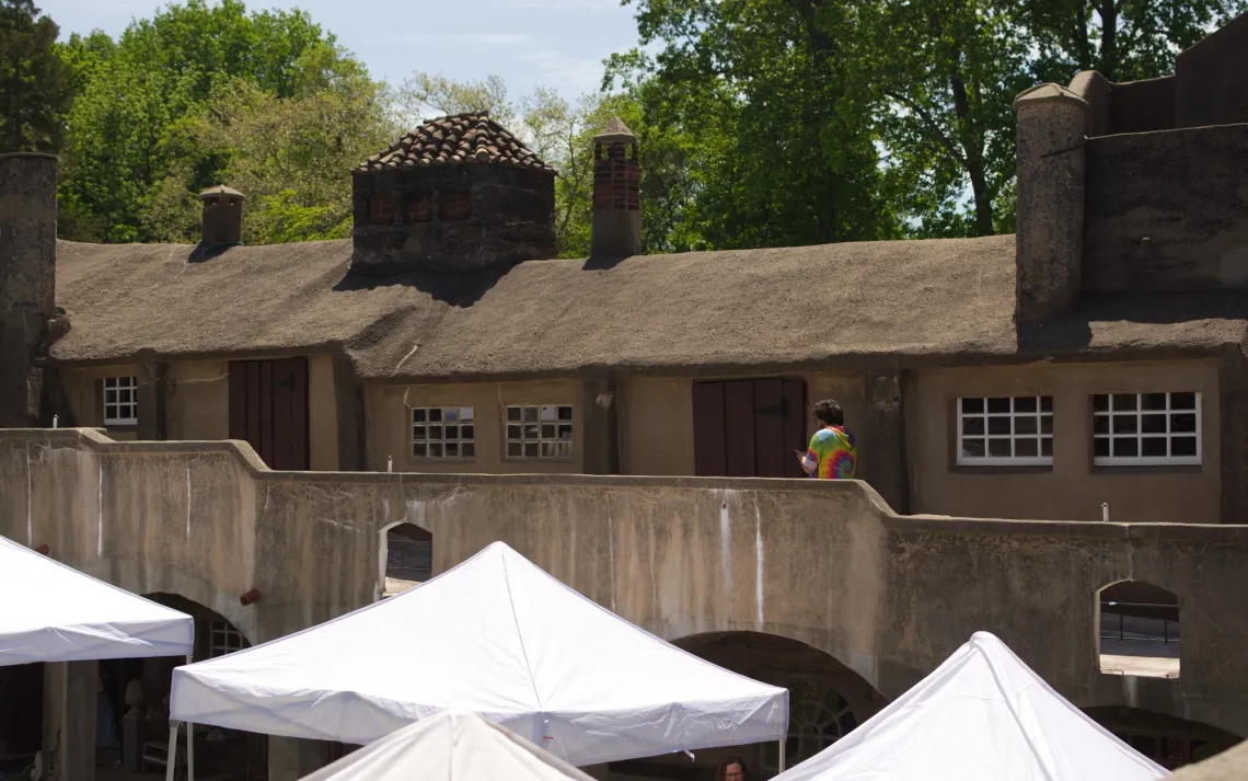 A person in rainbow attire walks across the upper balcony of a factory, with white tents in the foreground on the courtyard below.