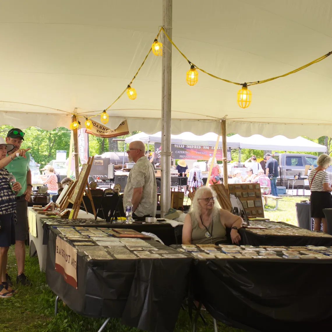 Under a white tent at an arts market, black table booths display tiles. Behind the booths, string lights are attached to metal poles that hold up the tent.