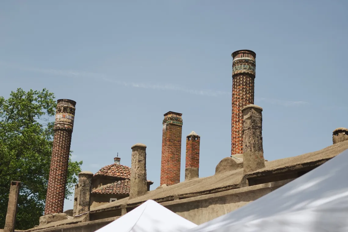 Brick chimneys of various sizes at a clay factory point up to a blue cloudless sky. White tents reflect sunlight in the foreground.