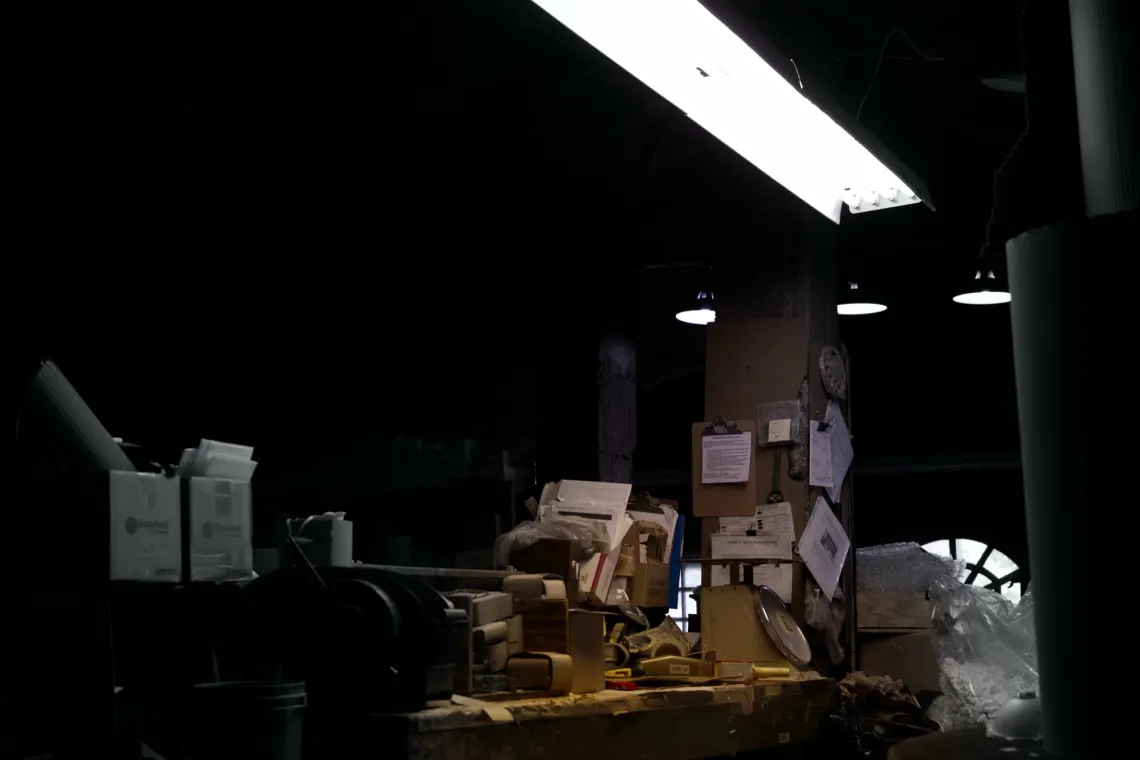  Workshop desk is lit by one overhead fluorescent light, in an otherwise dark room. The desk holds many boxes and papers.