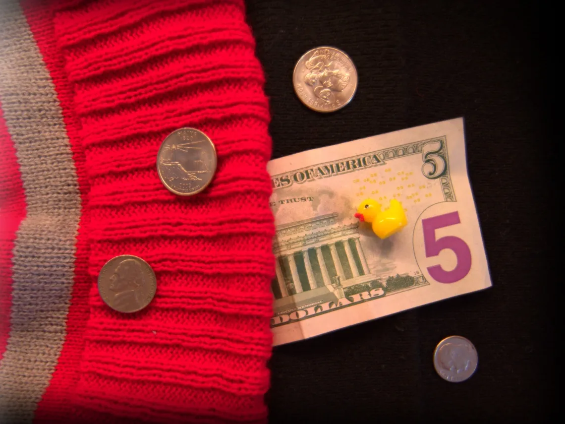 A five dollar US bill, various US coins, and a yellow plastic duck are surrounded by part of a red and gray beanie (left) and a black surface (right).