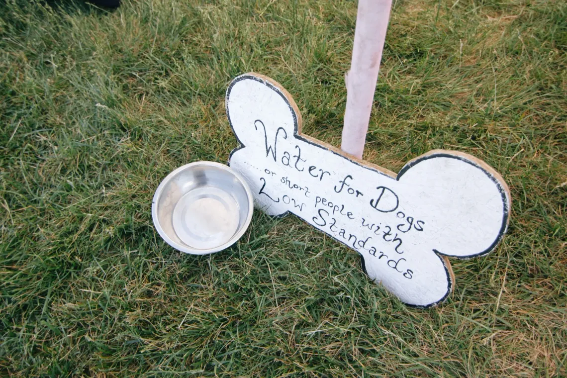 On the grass floor, a small metal dog bowl is empty. A sign next to it in the shape of a bone reads, "Water for Dogs ... or short people with Low Standards".