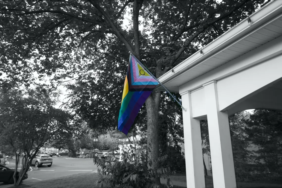 A 2021 Progress Pride flag points out from a white hut (right). The flag is the only object in color; everything else is in monochrome.