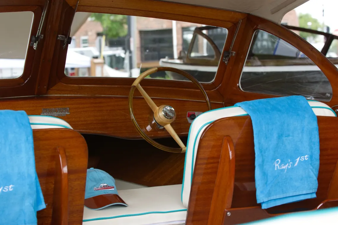 The front of a polished wooden boat, with steering wheel, is decorated with blue caps and towels that read "Ray's 1st".