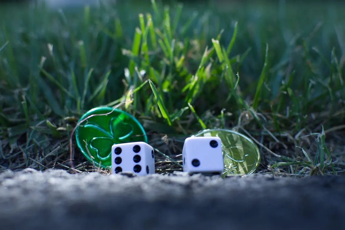 Two dice and two plastic coins are lit from above, with asphalt in the foreground and grass in the background.