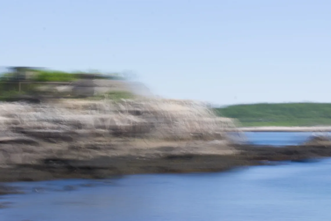 Horizontal motion blur image of a rocky shoreline over a blue cove of water. The sky is clear.