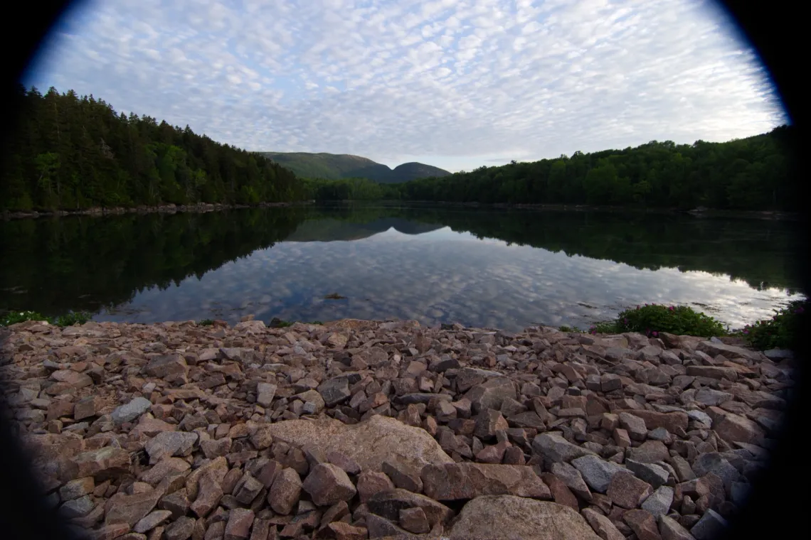 A rocky surface in the foreground meets with a body of still water. In the background, mountains and trees are reflected in this water.