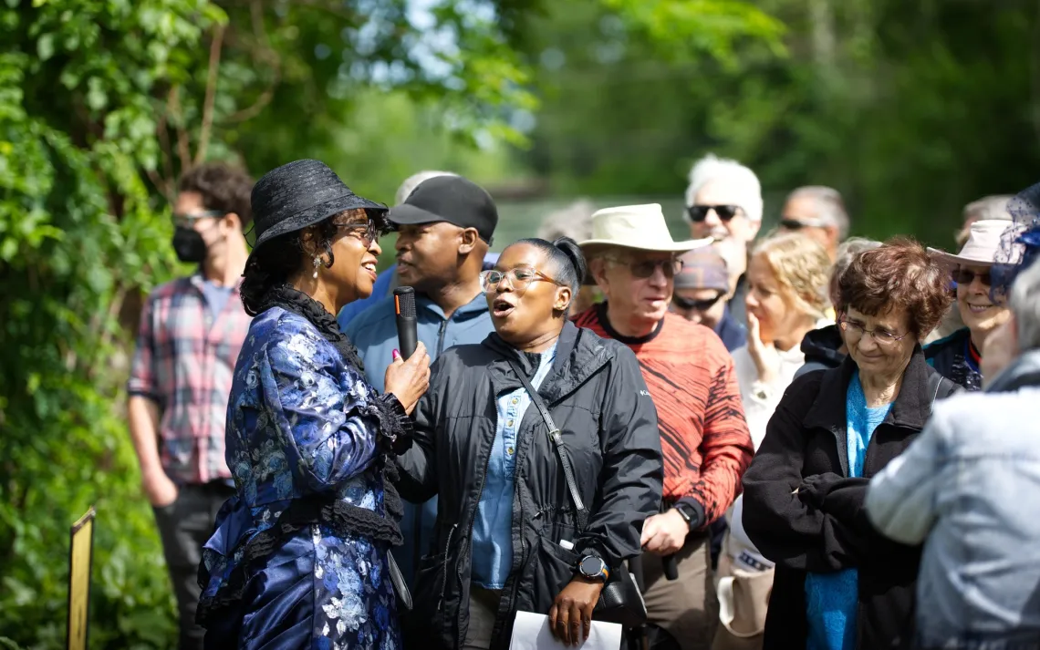 A woman in blue attire speaks into a microphone to a crowd. They are gathered on a dirt path next to trees and vines on a sunny day.