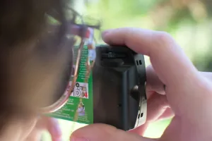Person with sunglasses holds a plastic film camera up to their eyes, looking at the greenery ahead.