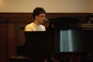 A boy with a white shirt sings into a microphone, sitting down in front of a black grand piano.