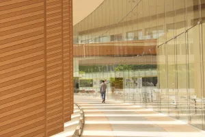 Lit by stripes of light through the orange louvers (left), a man in gray attire walks through an outdoor hallway with a curved glass wall to his right.