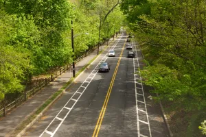 An overhead shot of a road with cars moving to and from the camera's point of view. The road is surrounded by a sidewalk (left) and trees.