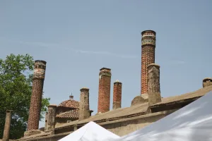 Brick chimneys of various sizes at a clay factory point up to a blue cloudless sky. White tents reflect sunlight in the foreground.