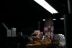  Workshop desk is lit by one overhead fluorescent light, in an otherwise dark room. The desk holds many boxes and papers.