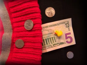 A five dollar US bill, various US coins, and a yellow plastic duck are surrounded by part of a red and gray beanie (left) and a black surface (right).