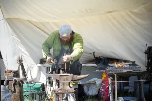 A man in green attire and dark apron marks a metal sheet on an anvil. He is surrounded by tools and an open fire (right).